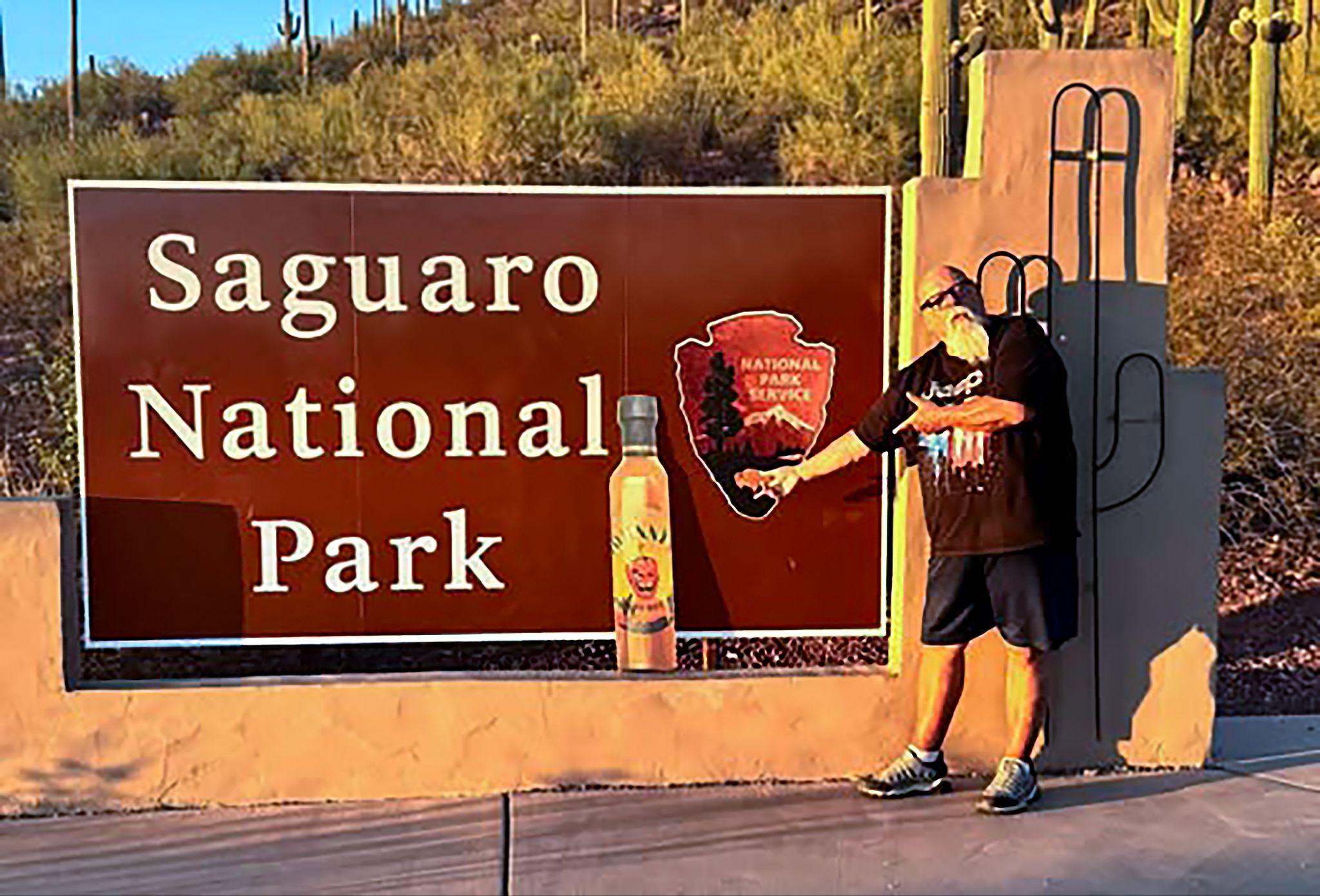 Johnny pointing at a bottle of Johnny's hot sauce in front of the Saguaro National Park entrance sign